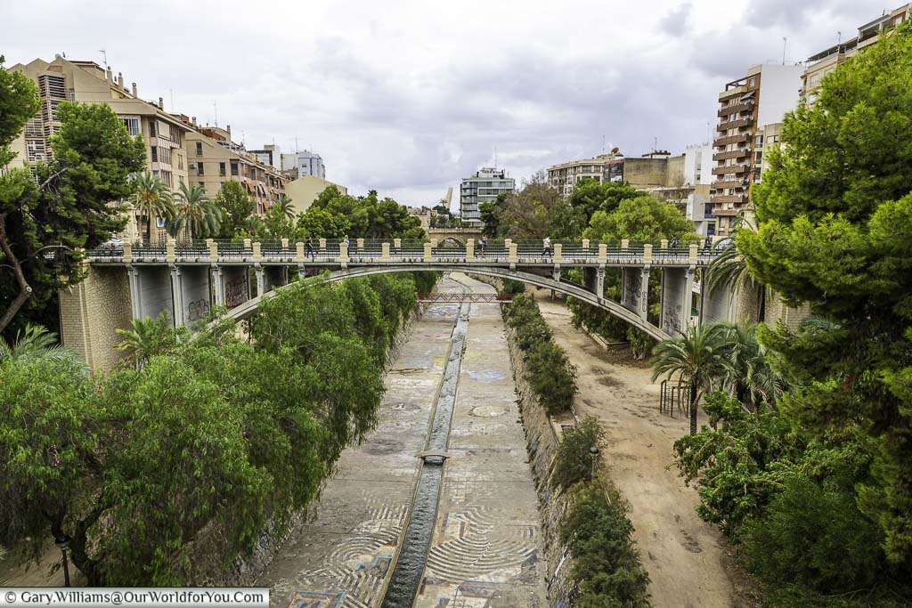 A photo of a pedestrian bridge in an art nouveau style crossing the River Vinalopó in Elche. Trees line the banks of the river, and buildings can be seen in the background.