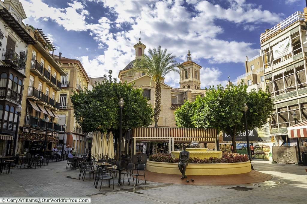 a fountain with a bronze statue of a seated lady in the centre of the Plaza de las Flores, surrounded by buildings on all sides, in murcia, spain