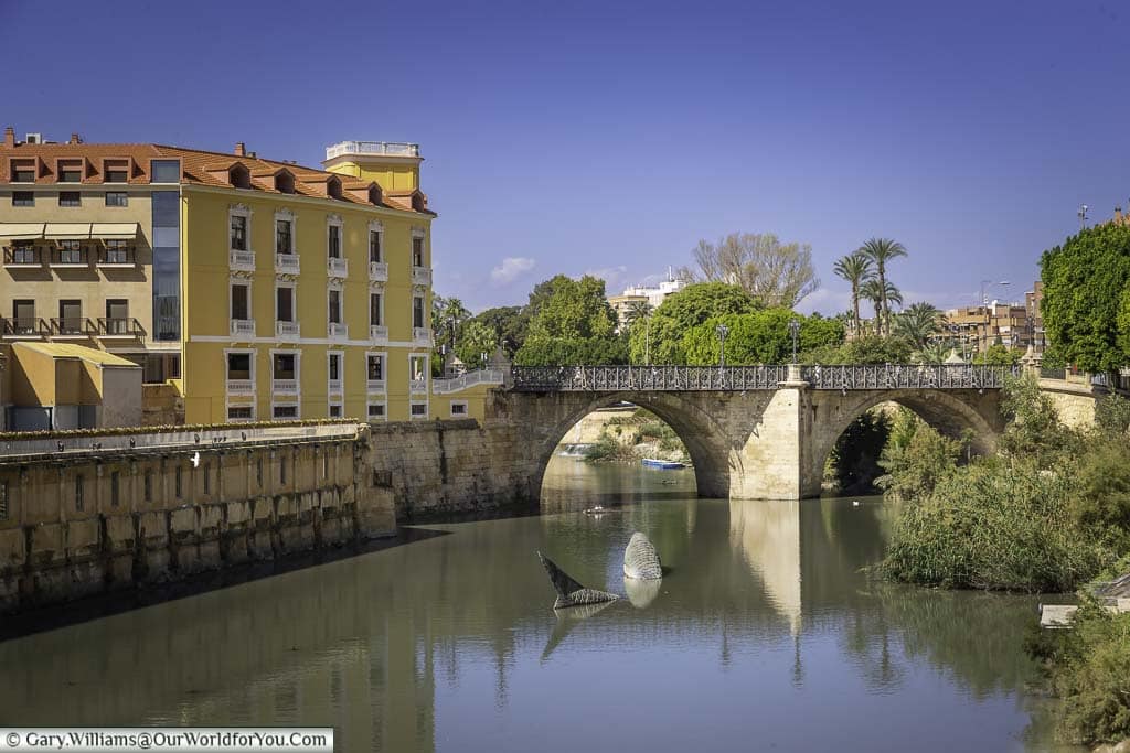 A view of the Puente de los Peligros, a bridge with multiple arches, spanning the Segura River in Murcia, Spain. In the distance, there is a statue of a whale breaching the water.