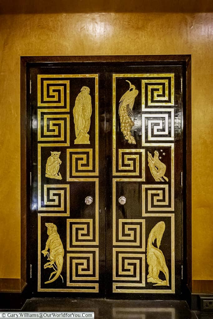 The black wooden door to eltham palace's dining room with gold geometric shapes and exotic animal inlays. The door is framed by wooden panelling and has a brass doorknob and hinges.