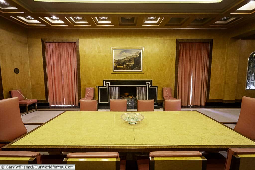 The dining room at eltham palace with a rectangular wooden table and ten art deco leather chairs set around it. The chairs have high backs and are arranged on a plain rug. In the background, a large art deco fireplace with a black surround is flanked by two windows with dark dusty pink curtains.