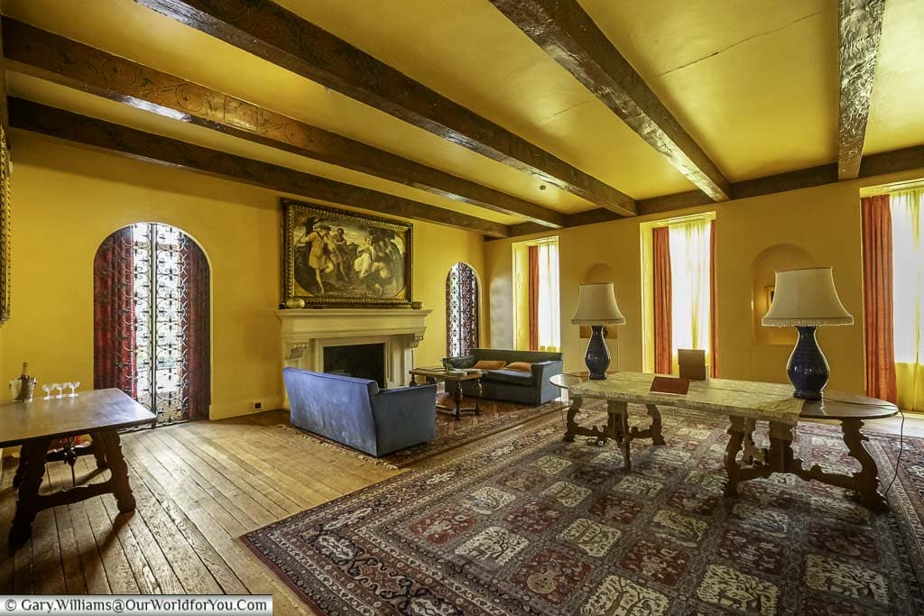 The drawing room at eltham palace, featuring a plush blue couch, a traditional fireplace, and a decorative table. The room has an elegant feel with high ceilings and heavy wooden beams.