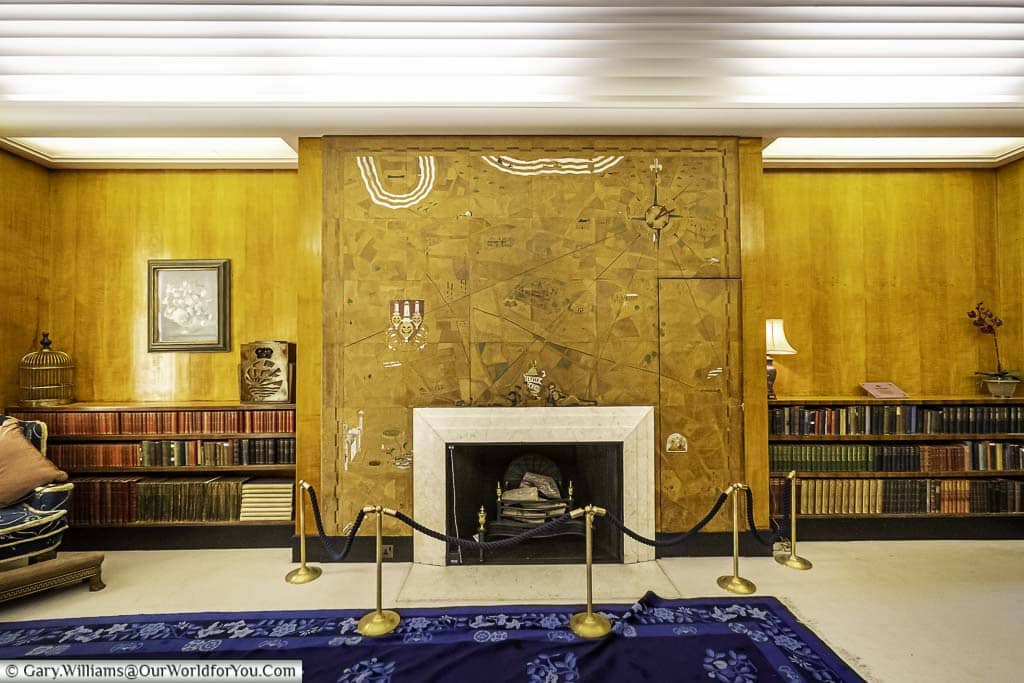 A photo of an art deco library with a large fireplace in the center, surrounded by short bookshelves filled with books. The bookshelves run a third of the height of the wall, and the chimney breast is decorated with map elements.
