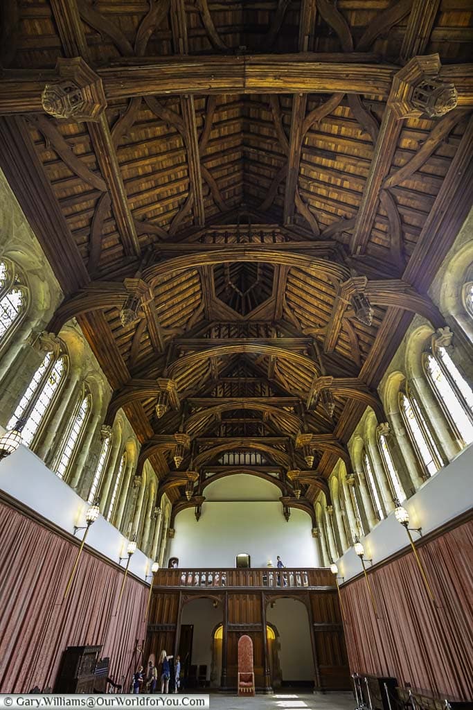 The richly detailed wooden ceiling of the grand hall at Eltham Palace with walls are lined with lanterns and dusty pink drapes.