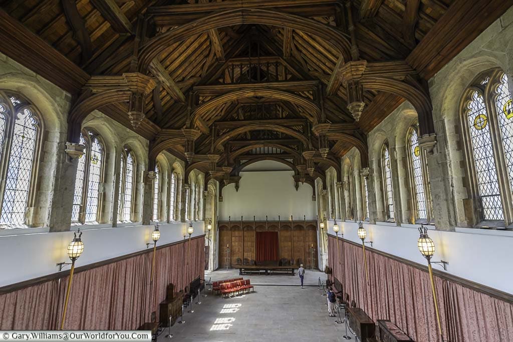 The interior of the grand hall at Eltham Palace with a richly detailed wooden ceiling and stained glass windows. The floor is polished stone, and the walls are lined with lanterns and dusty pink drapes.