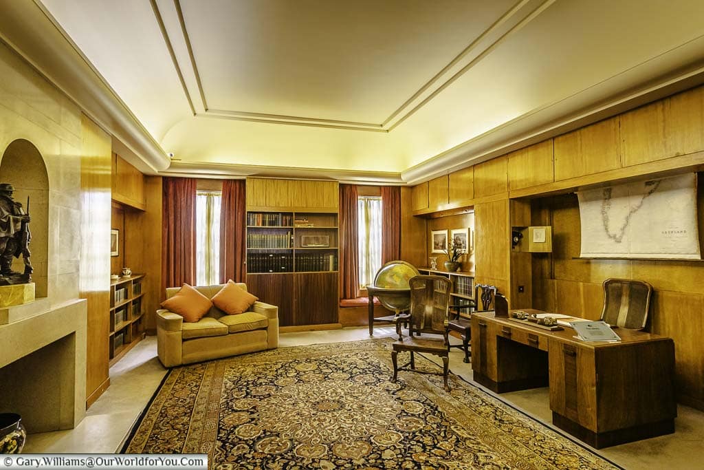 A photo of the library at eltham palace with a large, plush couch facing a wooden desk and chair. Sunlight streams through a window onto the desk, illuminating the library.