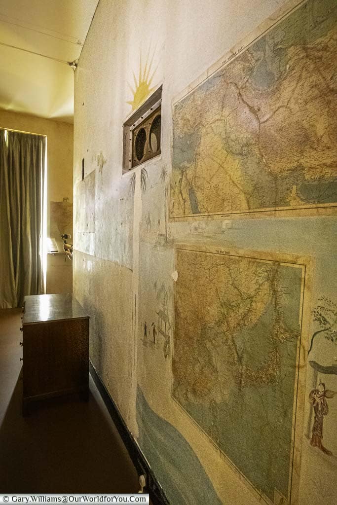 A narrow passage behind the library at Eltham Palace where the wall behind the desk is covered in framed maps, including a world map, a city map, and a nautical chart.