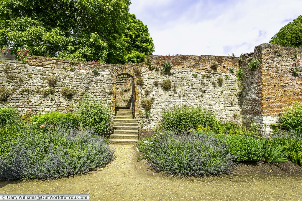 A close-up view of a stone wall and steps leading to a doorway in a garden of eltham palace. The wall is covered in moss and vines, and the steps are made of worn stone.