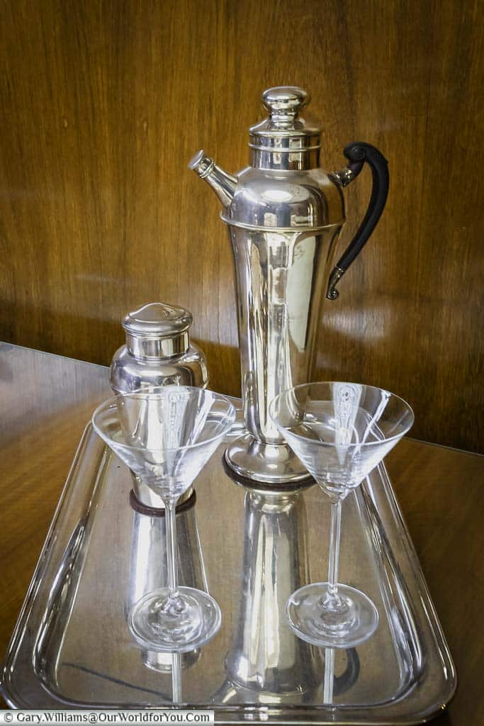 A silver cocktail shaker and two martini glasses on a black tray, sitting on a wooden table at eltham palace in south east london