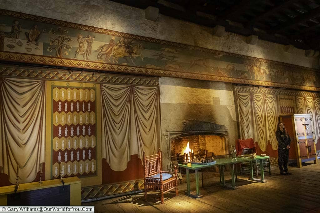 A fire roaring in a large stone fireplace inside dover castle keep. there is an english heritage guide next to a table in front of the fire, and the stone walls on either side are covered with tapestries.