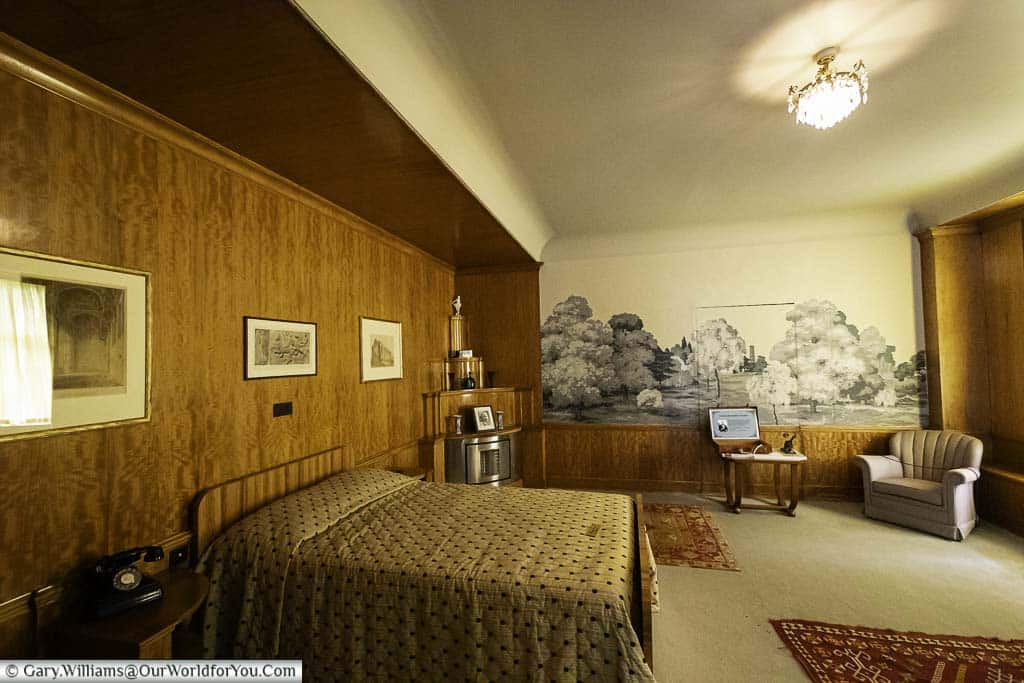A photo of a richly decorated bedroom in the art deco style at Eltham Palace, featuring a larger bed, a plush chair, a bedside table with a vintage telephone, and a painting. The bed has a gold bedspread in a geometric pattern.