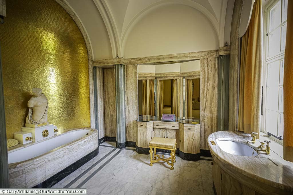 A brightly lit, opulent bathroom at Eltham Palace, with a large white bathtub, a pedestal sink, and a long vanity with a marble countertop. A white marble statue, possibly depicting a Roman deity, is displayed on the bathtub ledge.