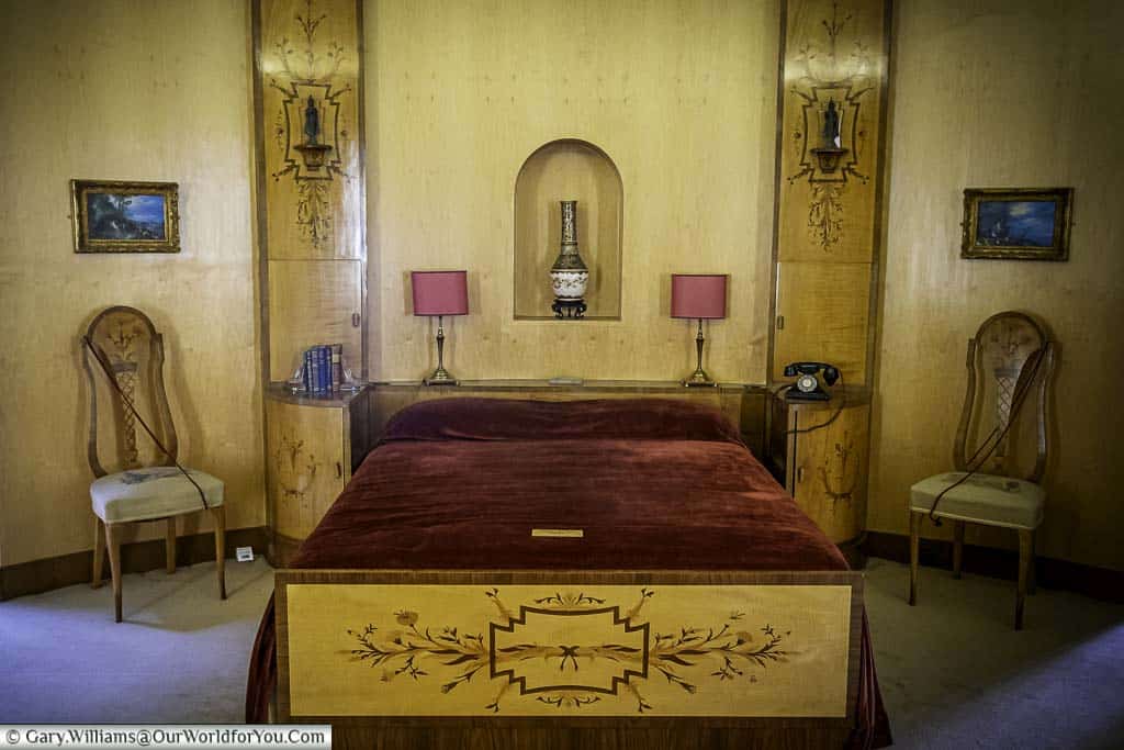 A close-up of a bedroom interior at Eltham Palace, London, showing a ornate art deco bed with a red and gold bedspread, two plush chairs, and two table lamps with patterned shades. A vintage telephone sits on a bedside table, and a large painting with ornate framing adorns the wall.