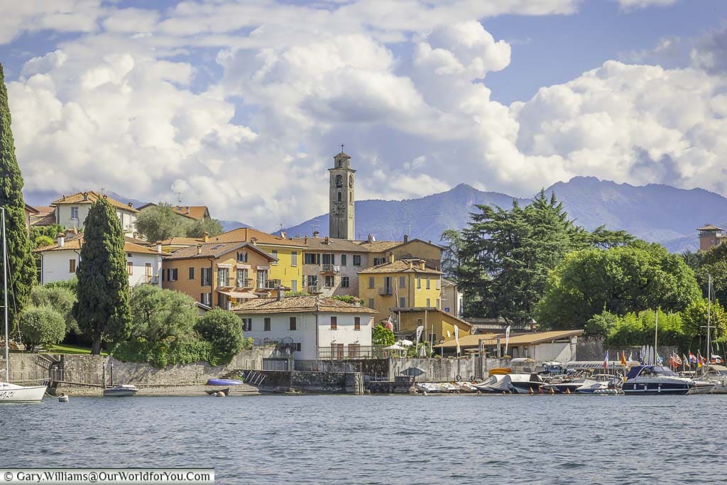 A town on the edge of Lake Como with small harbour at the front and the church tower dominating the skyline against the backdrop of mountains under a lightly cloudy sky.
