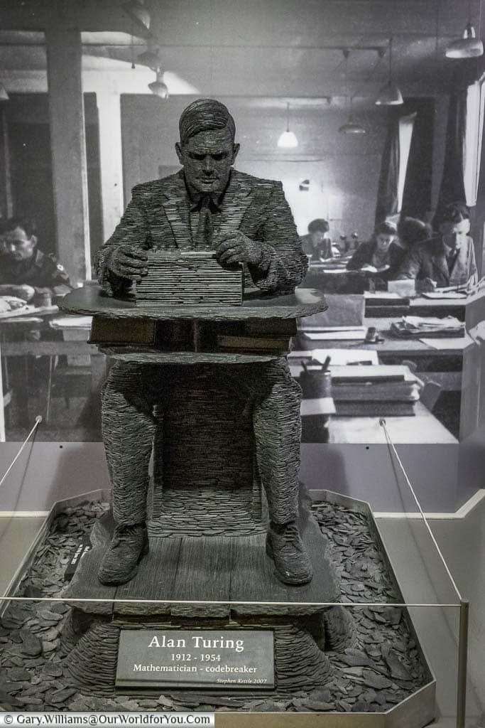 slate statue of a seated Alan Turing working on an Enigma machine, in front of a black & white image of the working interior of Block-B during the war.