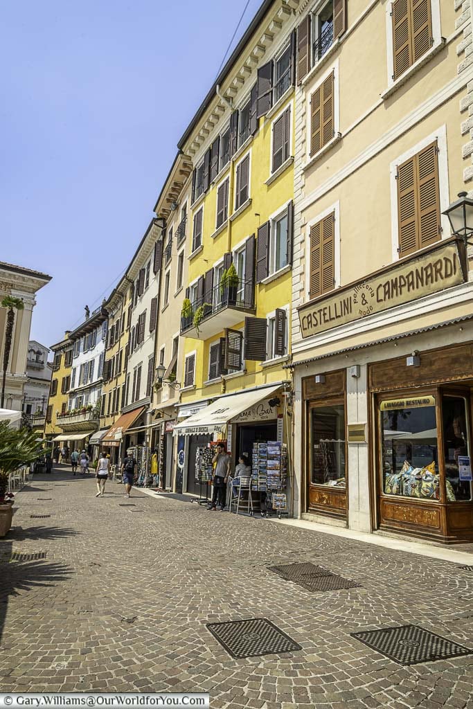 A cobbled lane that runs through the old centre of Salò. Lined on one side are tall, terraced buildings, with shops and restaurants in the base, and homes in the upper levels.