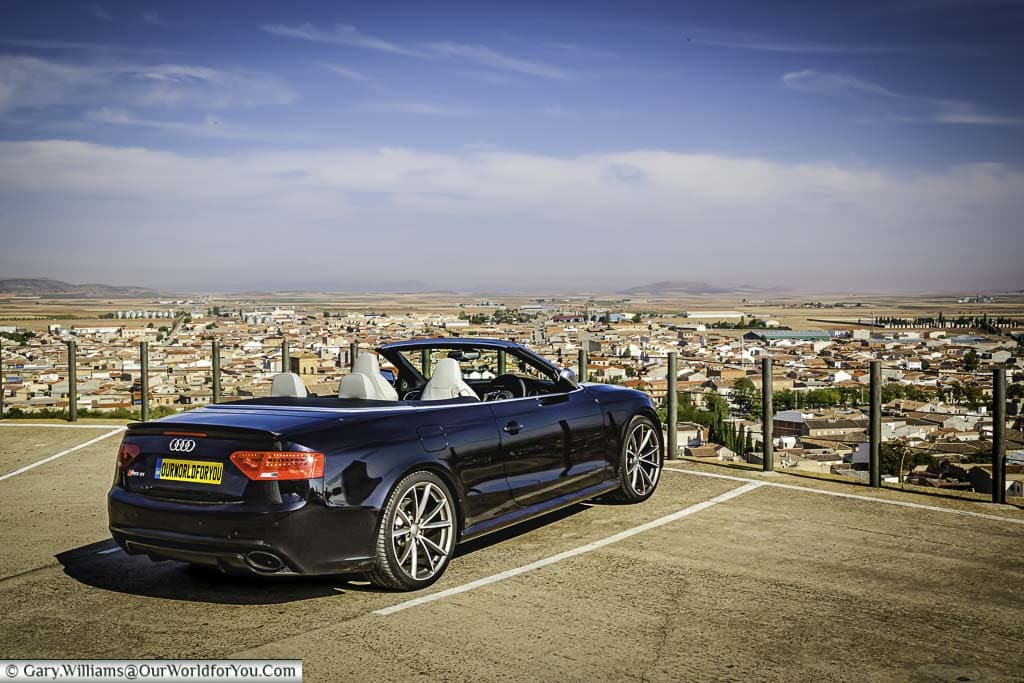 Our audi rs5 convertible, with the hood down, parked by the windmills of consuegra, overlooking the plains of la mancha in central spain on our spanish road trip
