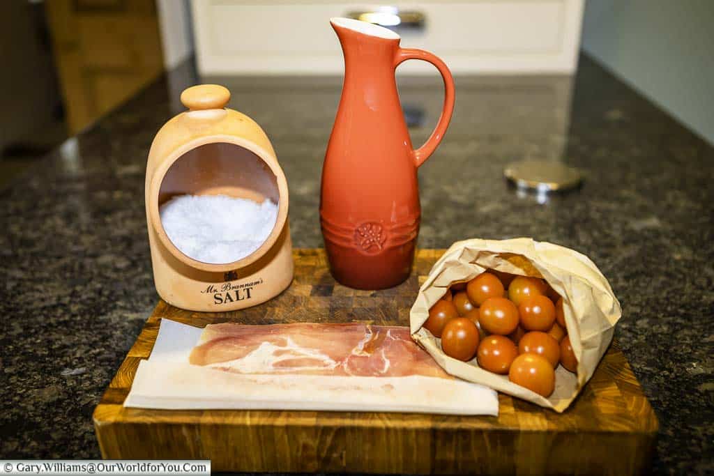 The basic ingredients for a Pan con Tomate, a salt pig filled with Maldon sea salt, a jug full of olive oil, a paper bag full of cherry tomates and some serrano ham wrapped in brown paper on a wooden chopping board.