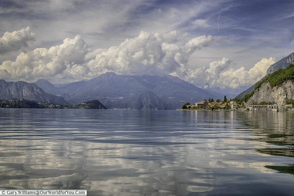 Shot taken from a boat on Lake Como where the light fluffy clouds in a blue sky are reflected in the water of the slightly rippling Lake. The vista is set against the mountainous backdrop and a small town on the right hand side edges into the Lake.