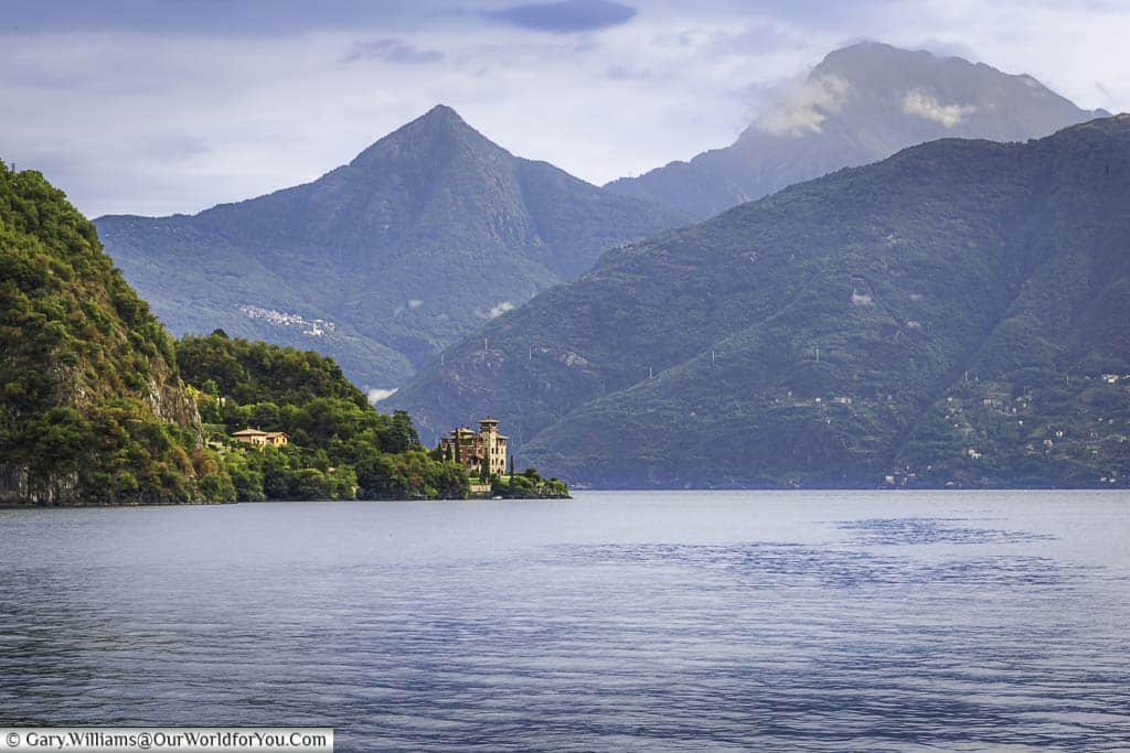 A shot taken from a boat on Lake Como of a villa at the edge of Lake Como with the green mountains acting as a backdrop against a slightly grey and cloudy sky.