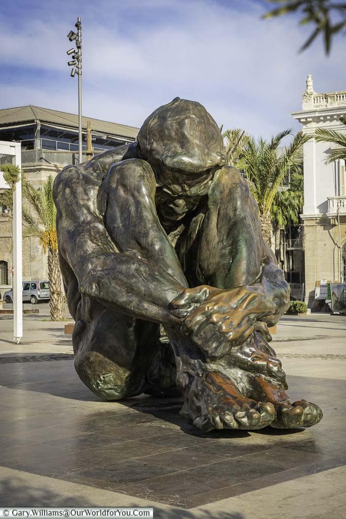 a bronze figure of a seated man, naked and helpless, hugging his legs and with his head down at the end of the esplanade of héroes de cavite in cartagena, spain